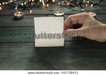 A photo of a hand holding a blank white thick cardboard business card next to a pile of them, on a dark wooden texture with candles in the background. A mockup or a banner with copyspace