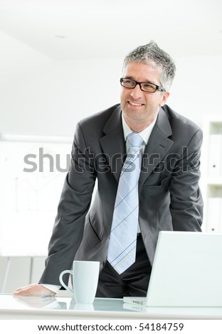 Gray haired businessman using laptop computer, standing behind office desk, working.
