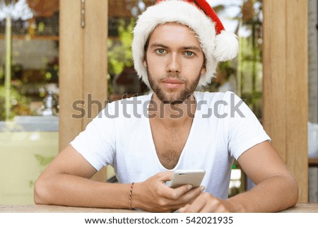 People, holidays, technology and communication. Young hipster with stylish beard using free wi-fi on cell phone, reading messages from his friends, congratulating him on Christmas via social networks