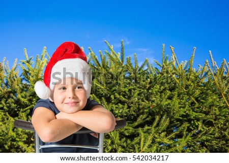 Handsome boy in a red cap of Santa Claus smiling. Background - green hedge firs