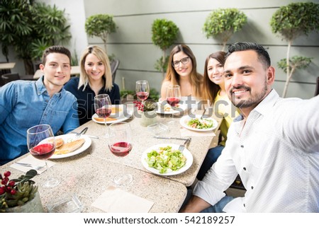 Wide view of a group of friends eating together in a restaurant and taking a selfie with a camera