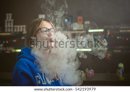 beautiful young woman with glasses smoking electronic cigarette. girl in a blue sweater blows clouds of glycerol vapor, from vape mod, sitting indoors vape shop cafe