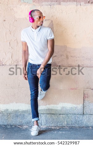 African American Man listening music in New York, wearing white Polo shirt, blue jeans, white sneakers, sunglasses, wireless headphones, standing against wall on street. Color filtered effect
