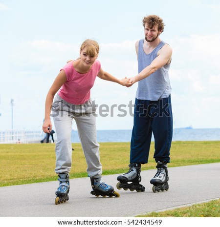 Active holidays, exercises, relationship concept. Young woman dressed in sports clothes putting her boyfriend up to do rollerblading while holding his hand on promenade