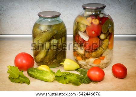 Home canning: large glass cylinders with a variety of vegetables: cabbage , tomatoes, cucumbers, peppers. Sealed with metal caps. Located next to fresh vegetables.