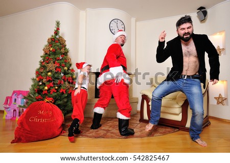 Good New Year spirit: Christmas tree, fireplace, big red gift bag - Santa and a hipster in corduroy jacket dancing and playing fool with a cute little boy in Santa's red cap, red pants and high socks