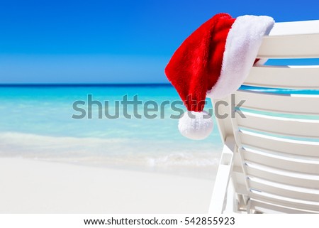 Santa Claus Hat on sunbed near tropical calm beach with turquoise caribbean sea water and white sand. Christmas vacation celebration Royalty-Free Stock Photo #542855923