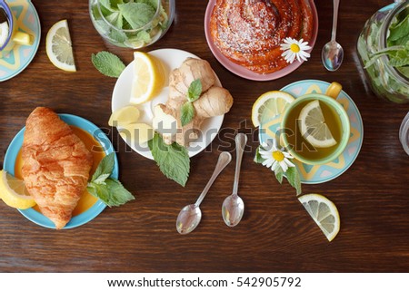 Cup of herb tea with lemon and mint leaves, ginger root and croissant on the wooden background