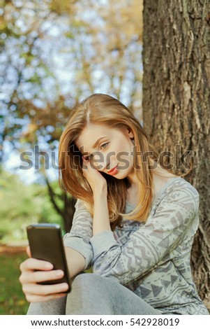 Beautiful girl with long blond hair and blue eyes sitting in the park and makes selfie. Location of autumn park.