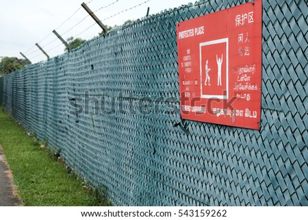 Barbed wire metal security fence with a red color warning sign for protected place in English, Chinese, Tamil and Malay language.