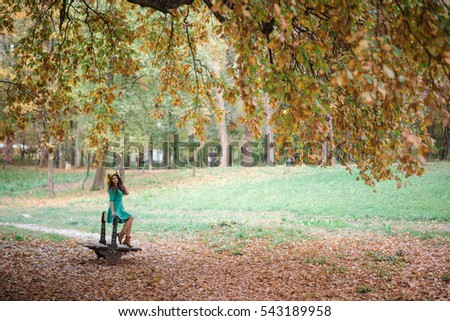 Beautiful woman in green with a yellow wreath posing in autumnal park. Young brunette woman spending time in the autumn of near a tree in the forest. Beautiful woman staying on the bench.