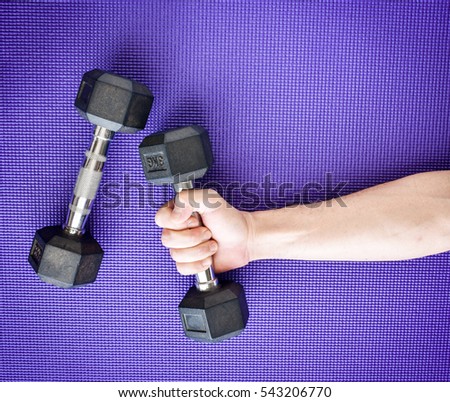 Workout equipment in fitness gym.Two dumbbells with one Asian man's hand lifting, purple blue yoga mat background.