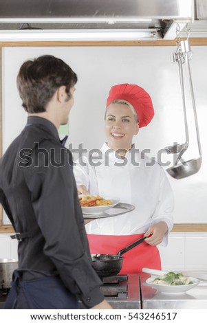 Male waiter receiving his next order from an attractive female cook in industrial kitchen wearing black uniform