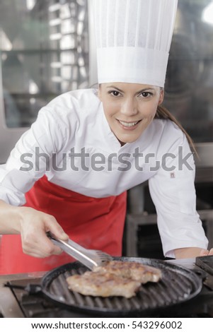 Young attractive female chef or cook in industrial kitchen brushing on some sauce on steak cooking on a cast iron grill