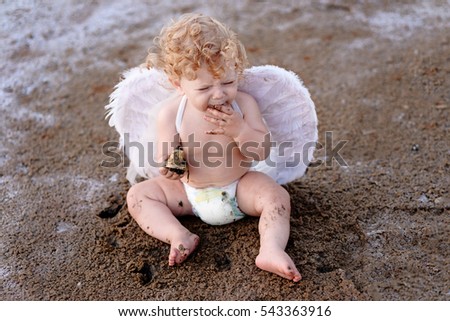 Dissatisfied little curly angel trying to taste the ground, sitting on the ground and playing with bell