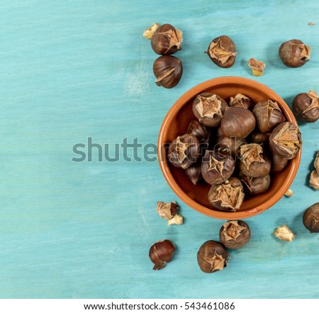 An overhead photo of peeled and unpeeled roasted chestnuts in an earthenware bowl, on a vibrant teal blue background with copyspace