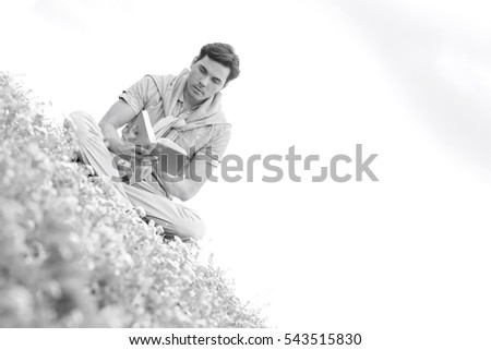 Tilt image of young man reading book while sitting on grass against sky
