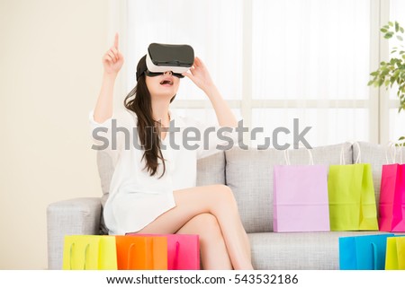 asian woman experience shopping online with VR headset sitting on sofa colorful shopping bags around. indoor living room background