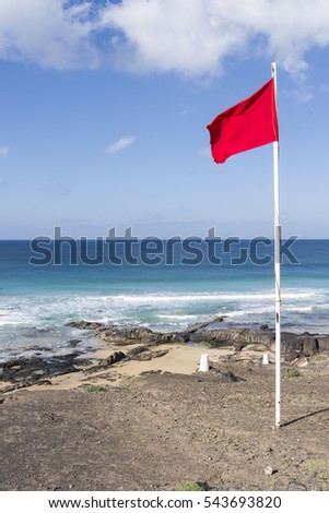 Red beach flag on a pole signaling alert pictured against deep blue sky. In the background the turquoise Atlantic Ocean with waves rolling in.
