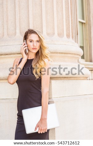 American Business Woman with long blonde hair working in New York, wearing black dress, holding laptop computer, standing by column on street, listening, talking on cell phone. Color filtered effect