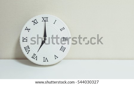 Closeup white clock for decorate show 7 a.m. on white wood desk and cream wallpaper textured background with copy space