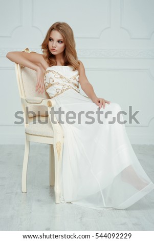 Smartly dressed pretty and elegant young girl (teenager) with frail figure and blond wavy hair wearing white evening dress embroidered with sequins is posing in the light interior studio room