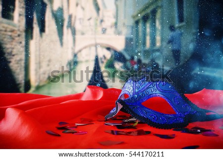 Image of elegant venetian mask on red silk fabric in front of blurry Venice background. Glitter overlay