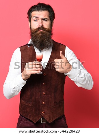 handsome bearded man with long beard and mustache on serious face holding glass of alcoholic shot in vintage suede leather waistcoat on red studio background