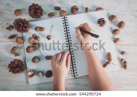 women write in open notebook with acorns and cones on white wooden background