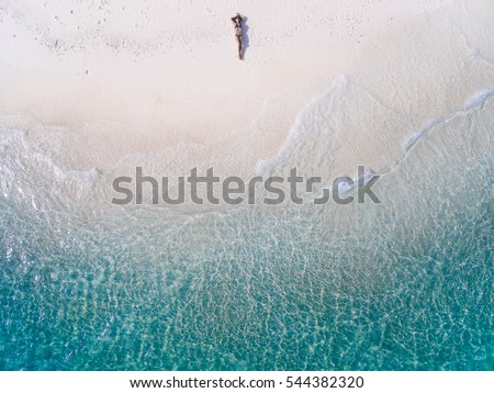 Young woman in a bikini lying on the back on the white sand near the waves of blue sea. Top view. Kai island, Andaman Sea, Phuket, Thailand. Aerial Shooting.