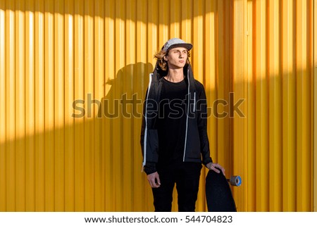 Young hipster guy portrait with longboard stranding at yellow wall