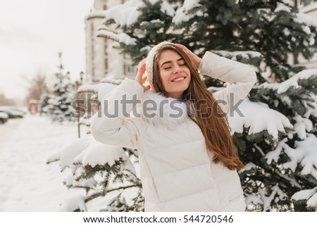 Portrait lovely cute girl chilling on sunshine in frozen morning. Young joyful woman enjoying winter time on fir trees full with snow background. Positive true emotions, smiling with closed eyes