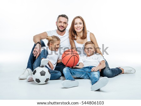 Cheerful family in white t-shirts and jeans sitting with soccer and basketball balls isolated on white
