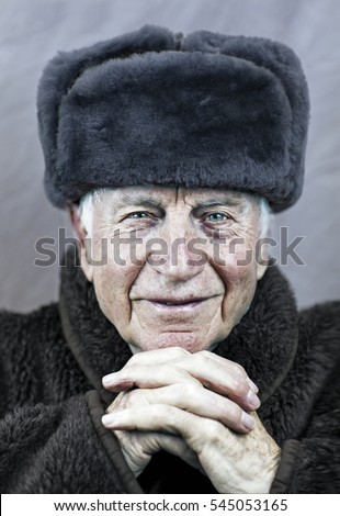 Old Caucasian man with wrinkled weathered face with vibrant blue eyes wearing a soviet era Russian military uniform with beaver fur winter hat with his hands crossed across his chin