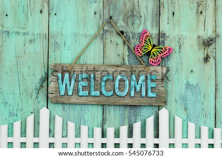 Welcome sign with butterfly hanging over white picket fence by antique rustic mint green wood background; colorful springtime composition with wooden painted copy space