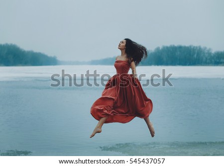 Beautiful brunette girl jumping, walking in the air. Background shore of a frozen river.  Sad face with a glimmer of hope to escape from the routine of everyday life. Fantasy photo, creative color.
