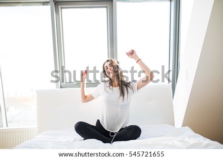 Happy woman or teenage girl in headphones listening to music from smartphone and dancing on bed at home