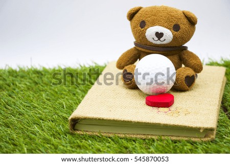 Bear is on art book with golf ball and red heart on grass.