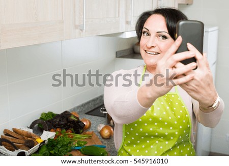Portrait of happy smiling russian housewife in apron making selfie in domestic kitchen