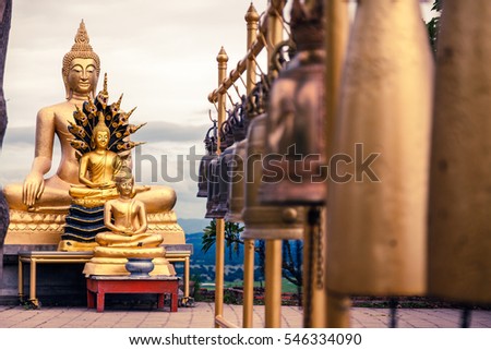 Three golden buddha statue sitting in line with many bells hanging.