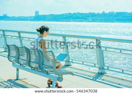 American Business Woman traveling, working in New York in hot summer, sitting on chair at park by Hudson River, facing New Jersey, reading at laptop computer. Back View. Color filtered effect
