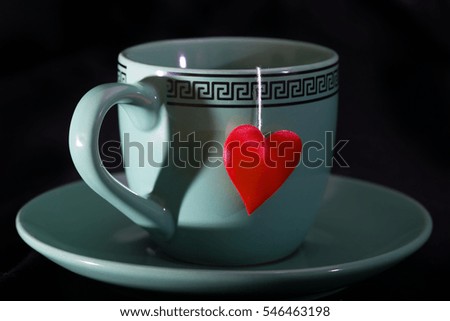 Cup with a shadow in the form of heart and a heart on a thread. A cup on a saucer, a red heart from a tea bag, a black background
