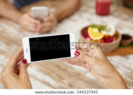 Female using online application for editing her pictures before posting them via social network while relaxing at coffee shop, sitting at wooden table with man holding gadget in blurred background