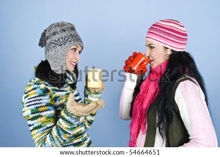 Two beautiful woman friends dressed in winter clothes enjoying a conversation,drinking hot drink and laughing together over blue background