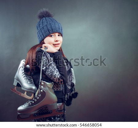 Cheerful little girl in warm sweater and hat holding figure skates.