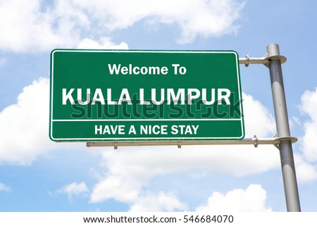 Green overhead road sign with a Welcome to Kuala Lumpur, Have a Nice Stay concept against a partly cloudy sky background.