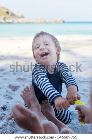 small adorable blond toddler playing with his sister, cheerfully laughing and looking at her, siblings sitting at beach together 