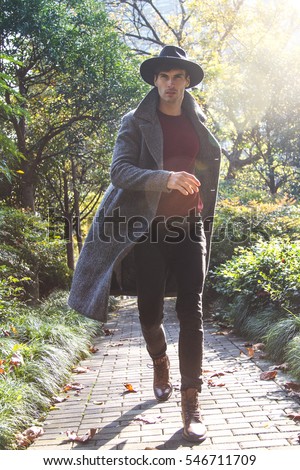 Handsome mature male model with beard, wearing black hat and long gray coat, he is walking in autumn garden and searching for the ray of sun. 