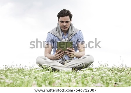 Full length of young man holding digital tablet while sitting on grass against sky