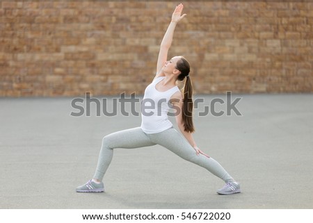 Sporty beautiful young woman practicing yoga, standing in Reverse Warrior exercise, Viparita Virabhadrasana pose, working out, wearing sportswear, outdoor full length, brick wall background